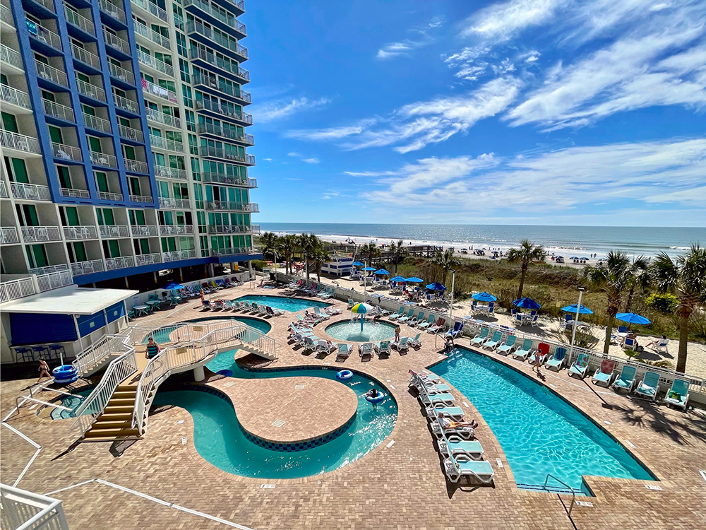 Outdoor pool complex with lazy river, swimming pool, pool chairs and beach umbrellas with a view of the ocean in the background
