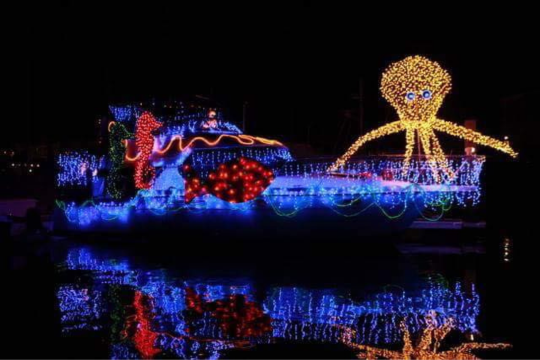 Boat adorned with festive lights and a large octopus figure reflected on water.