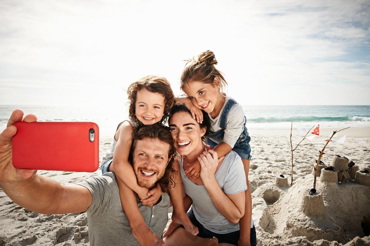 family with a mom, dad, son and daughter on the beach with the ocean in the background and a sandcastle they built taking a selfie together