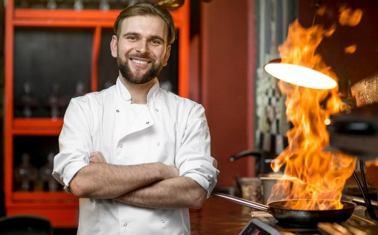 Smiling chef with arms crossed next to a flaming pan.