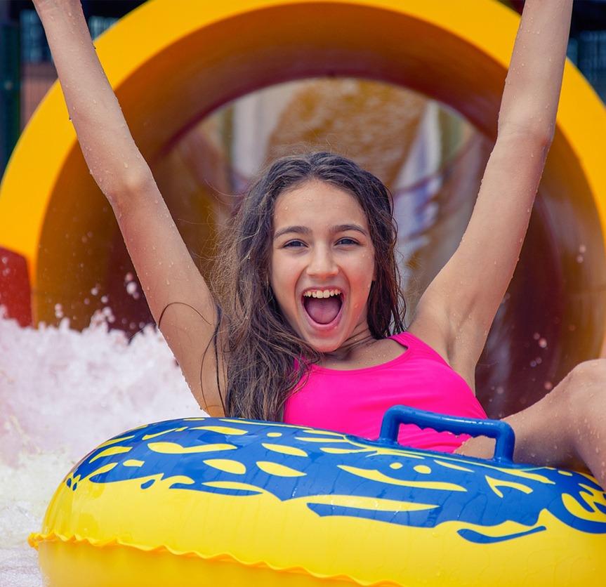 Young girl in a yellow innertube with her hands up exiting a yellow waterslide
