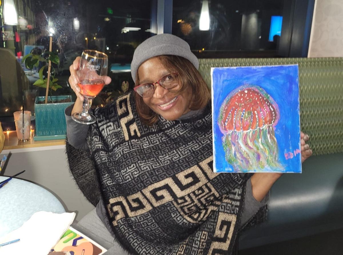 A woman holding up a handmade painting with a glass of wine